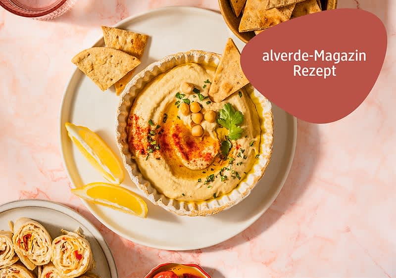 Teller mit Hummus garniert mit Kichererbsen, Petersilie und Paprikapulver, dazu Zitronenspalten und Tortillachips; rote Einblendung mit Text „alverde-Magazin Rezept“