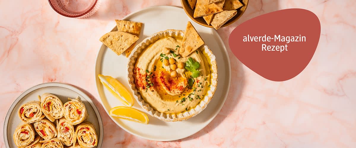 Teller mit Hummus garniert mit Kichererbsen, Petersilie und Paprikapulver, dazu Zitronenspalten und Tortillachips; rote Einblendung mit Text „alverde-Magazin Rezept“