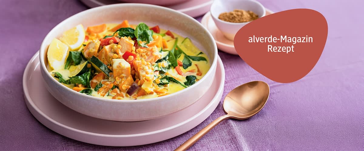 Schale mit cremiger Linsensuppe mit Spinat, Zucchiniwürfeln, roten Paprikastücken und Zitronenschnitz auf rosa Tellern, rechts oben roter Aufkleber mit weißer Schrift „alverde-Magazin Rezept“