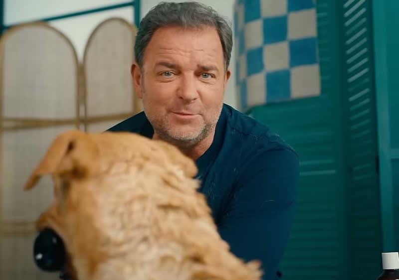 Ein Hund mit hellem Fell sitzt vor Martin Rütter, dessen Gesicht unscharf ist. Im Hintergrund sind grüne und beige Elemente zu sehen.