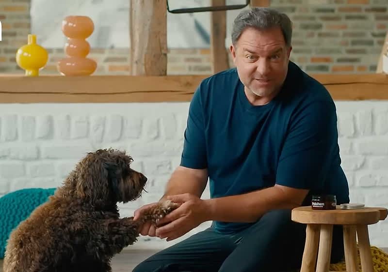 Martin Rütter in einem dunkelblauen T-Shirt hält die Pfote eines braunen, lockigen Hundes. Neben ihnen steht ein kleiner Holztisch mit einer Dose darauf. Im Hintergrund sind eine weiße Wand und dekorative Vasen zu sehen.