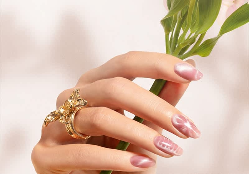 Eine Hand mit rosa glänzend lackierten Fingernägeln hält eine grüne Pflanze. Am Ringfinger befindet sich ein goldener Ring mit einem floralen Design.
