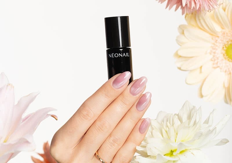 Eine Hand mit rosa lackierten Nägeln hält eine schwarze Flasche mit der Aufschrift „NEONAIL“, umgeben von verschiedenen Blumen in Pastellfarben