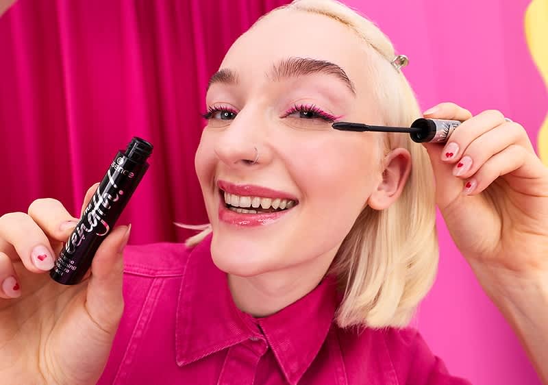 Eine Person mit blondem Haar trägt Mascara auf. Sie hält eine schwarze Mascara-Flasche in der Hand. Im Hintergrund ist ein pinker Vorhang zu sehen.
