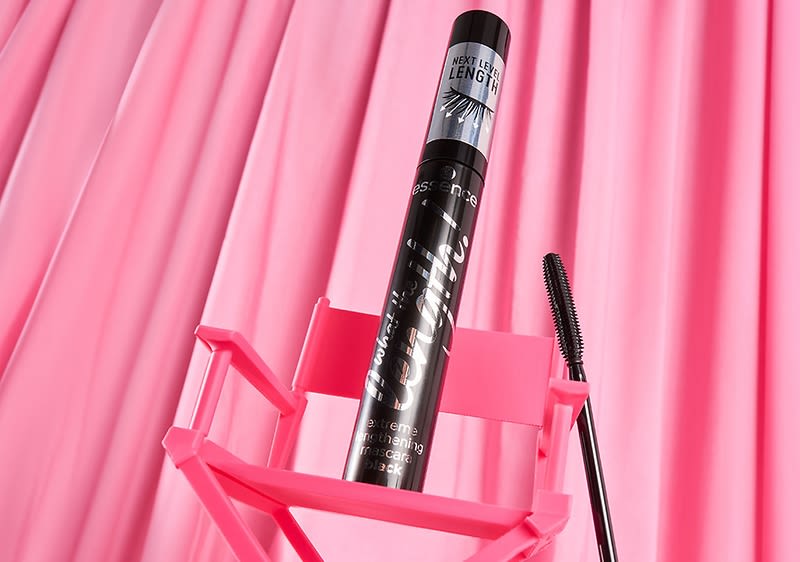 Eine schwarze Mascara-Flasche mit der Aufschrift „essence what the fake! extreme lengthening mascara black“ steht in einem pinken Halter vor einem pinken Hintergrund. Daneben liegt eine Mascara-Bürste.