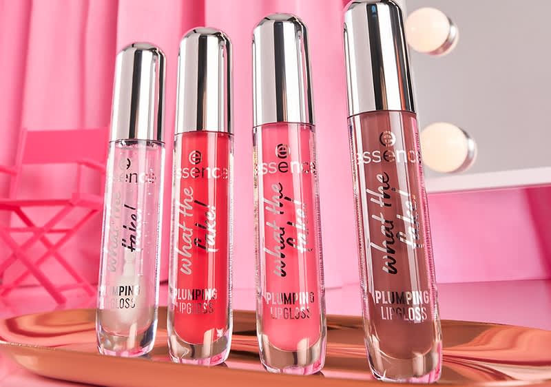 Vier Lipgloss-Flaschen von Essence stehen nebeneinander auf einem Tisch. Die Farben sind transparent, rot, rosa und braun. Im Hintergrund sind ein Spiegel und ein roter Stuhl vor einem rosa Vorhang zu sehen.