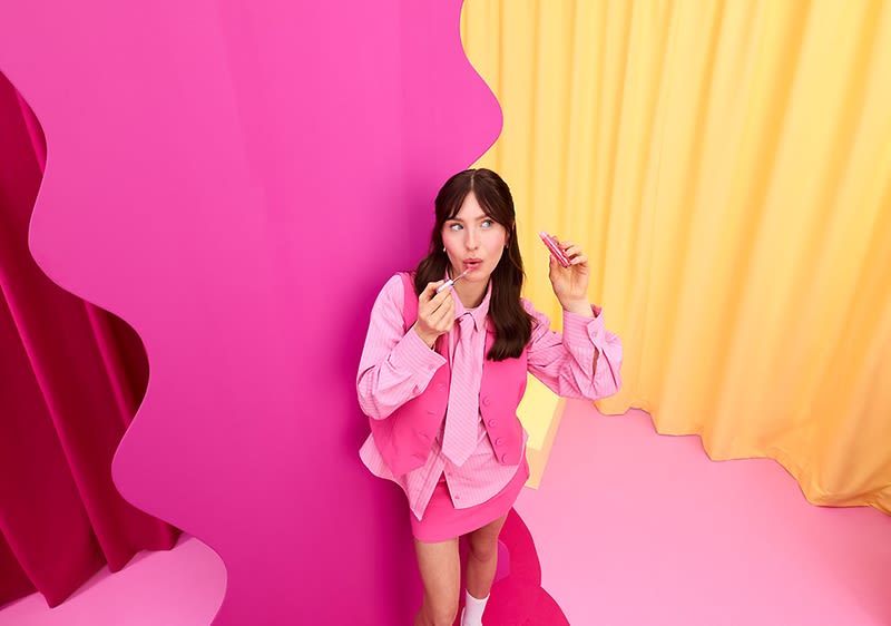 Eine Person in einem pinkfarbenen Outfit steht vor einem pinken und gelben Hintergrund und hält einen Lippenstift in der Hand