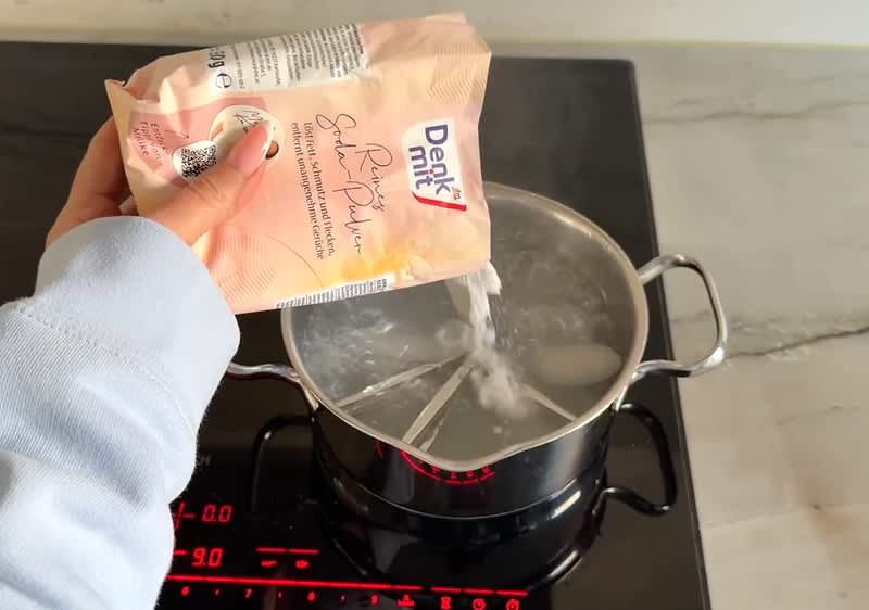 Eine Hand hält eine Packung Denkmit Spülmaschinen-Salz über einen Topf mit kochendem Wasser auf einem Herd mit digitaler Anzeige.