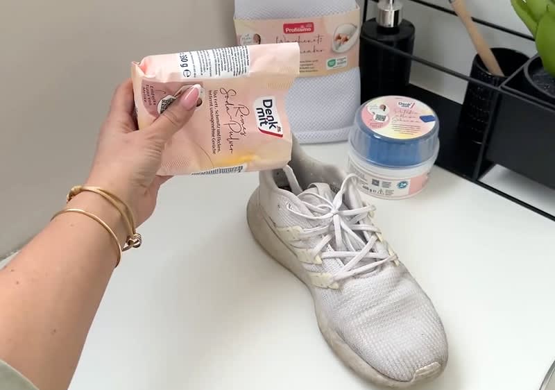 Eine Hand hält eine Packung „denk mit“ Seife, daneben ein weißer Turnschuh auf einem Tisch. Im Hintergrund sind weitere „denk mit“ Produkte zu sehen.