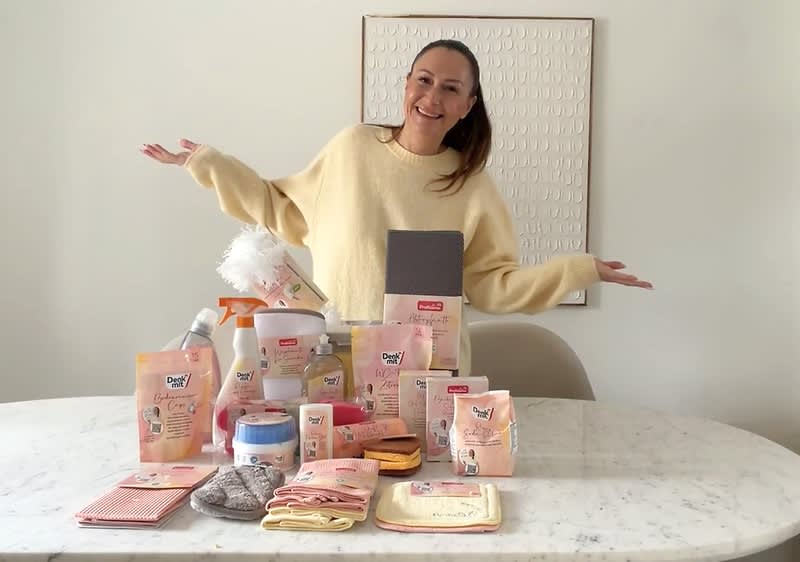 Eine Person steht hinter einem Tisch, auf dem verschiedene Babyprodukte der Marke Hipp angeordnet sind, darunter Feuchttücher, Cremes, Waschmittel und Pflegeprodukte. Die Person trägt einen hellen Pullover und hebt die Arme leicht an.