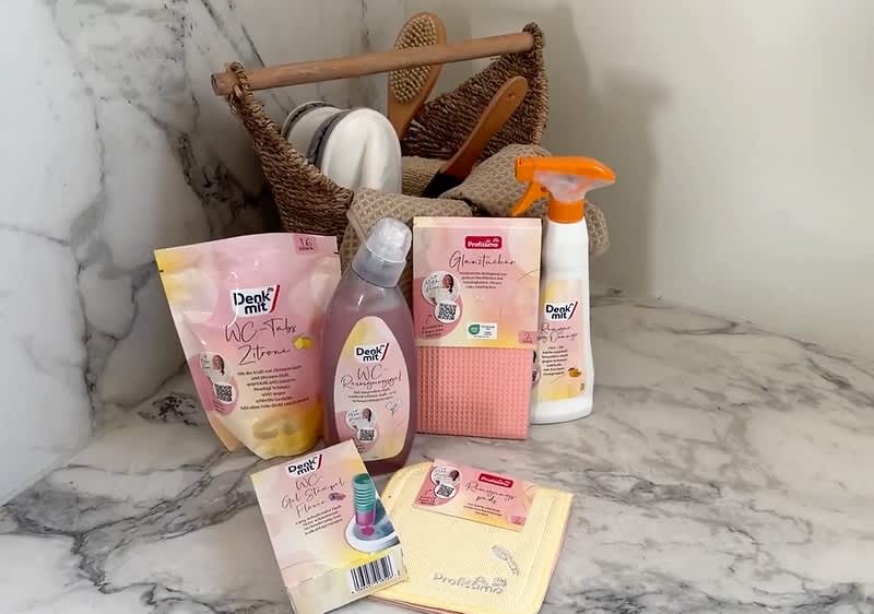 Eine Sammlung von Reinigungsprodukten der Marke „Dasch me“ auf einem Marmortisch. Zu sehen sind ein WC-Reiniger, ein Sprühreiniger, Reinigungstücher und ein Spülschwamm. Im Hintergrund steht ein Korb mit Reinigungsutensilien.