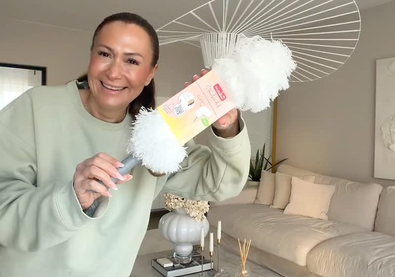Eine Person hält einen weißen Staubwedel mit einer Verpackung, auf der ein rosa und gelbes Etikett zu sehen ist, in einem modern eingerichteten Wohnzimmer mit beigen Möbeln und einem großen weißen Lampenschirm im Hintergrund