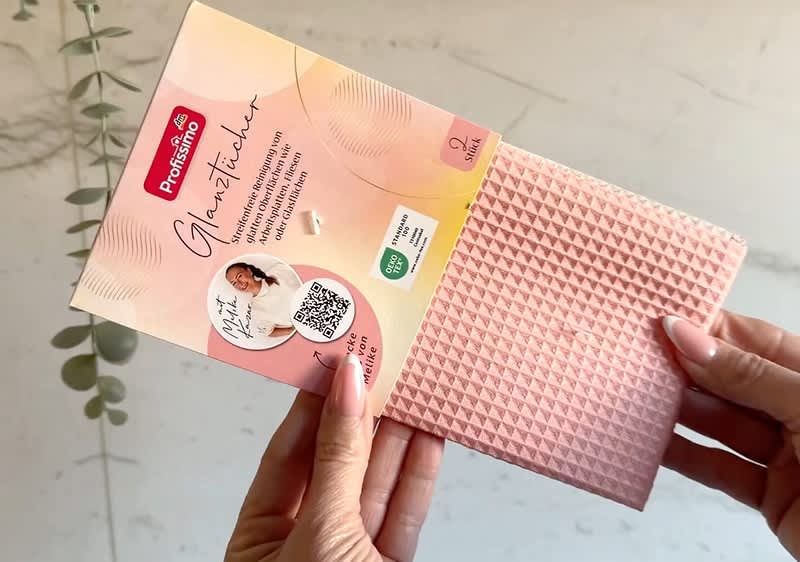Eine Person hält ein rosa Reinigungstuch mit einer Verpackung, auf der „Glanzstückchen“ steht. Die Verpackung zeigt ein Foto einer Frau, einen QR-Code und das Logo von Profissimo. Im Hintergrund ist ein Eukalyptuszweig sichtbar.