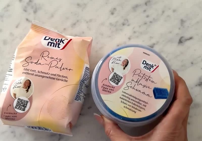 Eine Packung Denkmit Reines Soda-Pulver mit rosa und weißem Design und eine Dose Denkmit Putzhilfe mit Schwamm. Beide Verpackungen zeigen QR-Codes und Bilder von Personen.