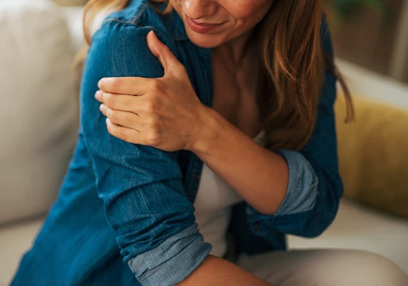 Eine Frau in einem blauen Hemd hält sich mit schmerzverzerrtem Gesicht die Schulter