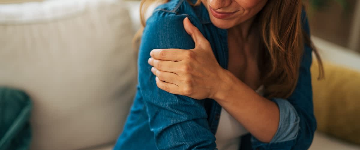 Eine Frau in einem blauen Hemd hält sich mit schmerzverzerrtem Gesicht die Schulter