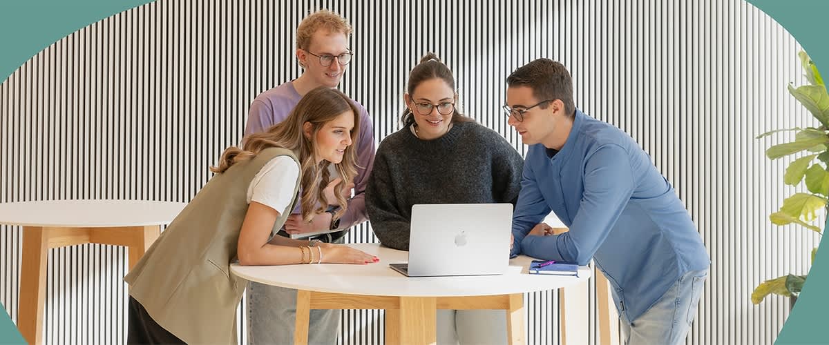 Vier Personen stehen um einen Tisch mit einem Laptop und besprechen Themen. Die Umgebung ist modern gestaltet.