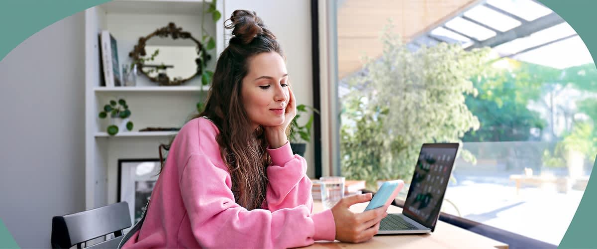 Eine Person in einem rosa Pullover sitzt an einem Tisch und hält ein Smartphone in der Hand. Neben ihr steht ein Laptop. Im Hintergrund sind Pflanzen und ein runder Spiegel zu sehen.
