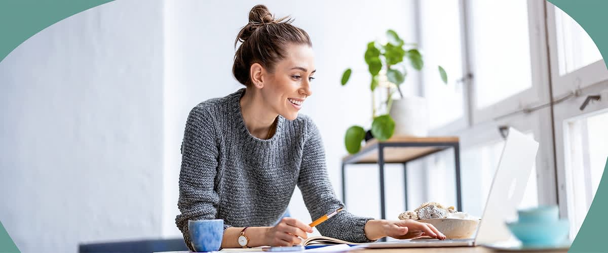 Eine Person in einem grauen Pullover arbeitet an einem Laptop, während sie einen Stift hält. Neben ihr steht eine blaue Tasse und im Hintergrund ist eine Pflanze auf einem Regal zu sehen.