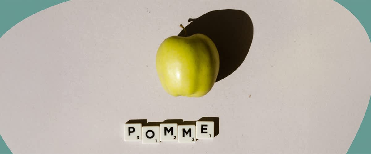 Ein grüner Apfel liegt auf einer Oberfläche, darunter sind Scrabble-Steine mit dem Wort „POMME“ angeordnet