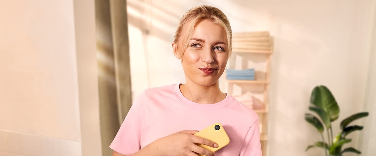 Frau mittleren Alters in rosa T‑Shirt hält ein gelbes Smartphone an die Brust und blickt nach rechts oben, im Hintergrund ein Regal mit Handtüchern und eine Zimmerpflanze