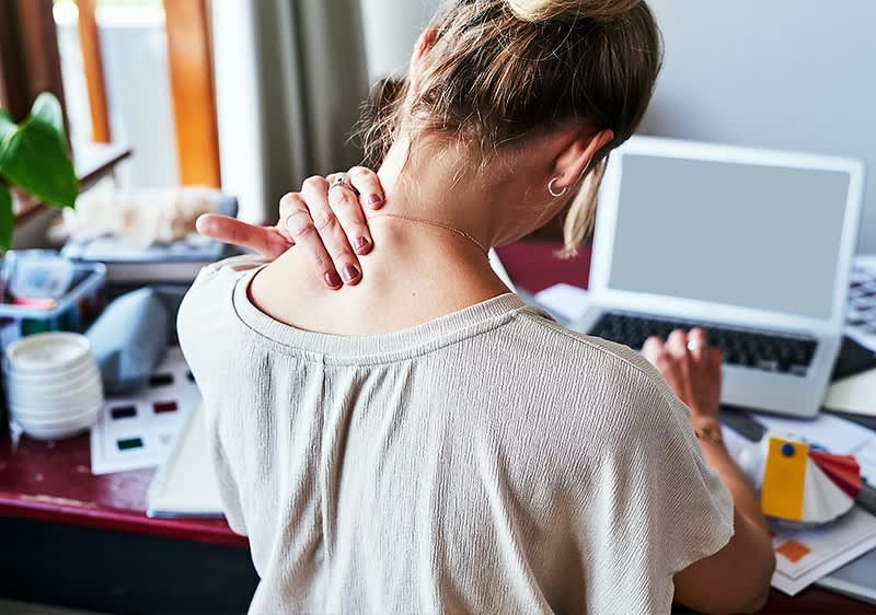 Eine Frau sitzt an einem Tisch vor einem Laptop und massiert ihren Nacken mit der linken Hand