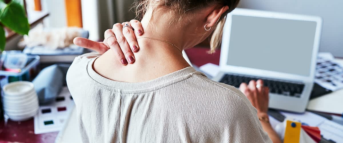 Eine Frau sitzt an einem Tisch vor einem Laptop und massiert ihren Nacken mit der linken Hand