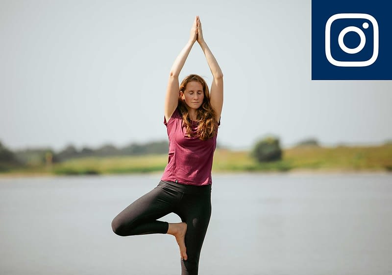 Eine Person steht in der Baum-Pose am Wasser, trägt ein lila Oberteil und schwarze Leggings. Oben rechts ist das Instagram-Logo zu sehen.