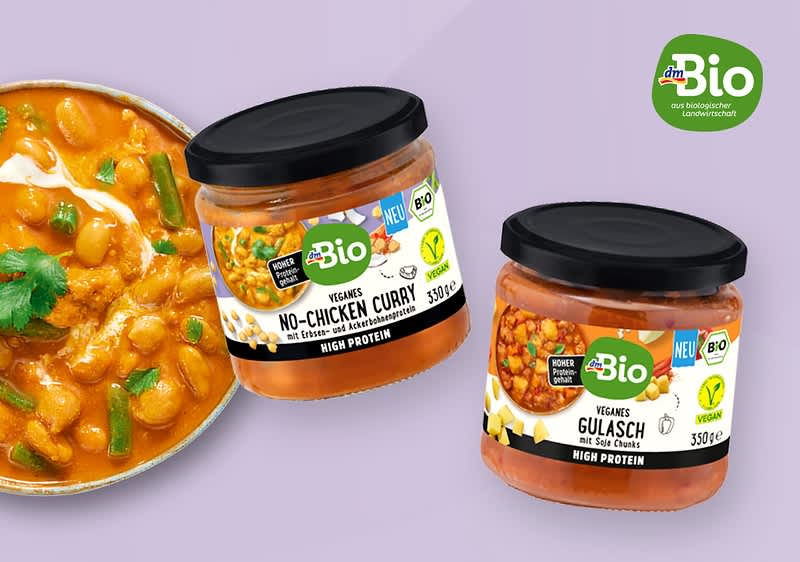 Zwei Glasgläser mit schwarzem Deckel und grüner „Bio“-Marke, links ein Glas mit Aufschrift „Veggans No‑Chicken Curry“ und Bild von Currystücken, rechts ein Glas mit Aufschrift „Veganes Gulasch“ sowie rechts oben grünes Bio-Logo, links daneben eine Schale mit orangefarbenem Currygericht garniert mit Korianderblättern