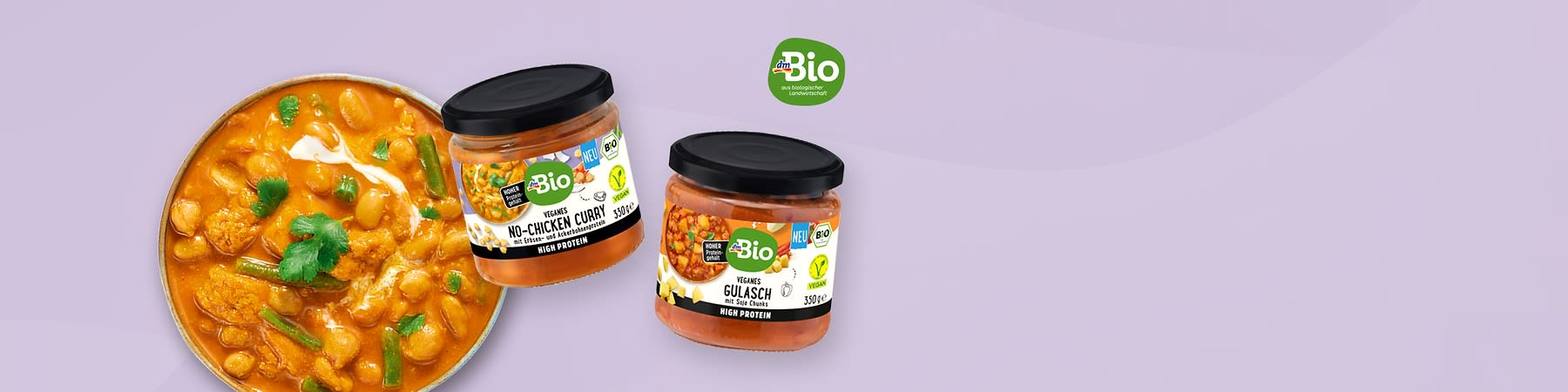 Zwei Glasgläser mit schwarzem Deckel und grüner „Bio“-Marke, links ein Glas mit Aufschrift „Veggans No‑Chicken Curry“ und Bild von Currystücken, rechts ein Glas mit Aufschrift „Veganes Gulasch“ sowie rechts oben grünes Bio-Logo, links daneben eine Schale mit orangefarbenem Currygericht garniert mit Korianderblättern