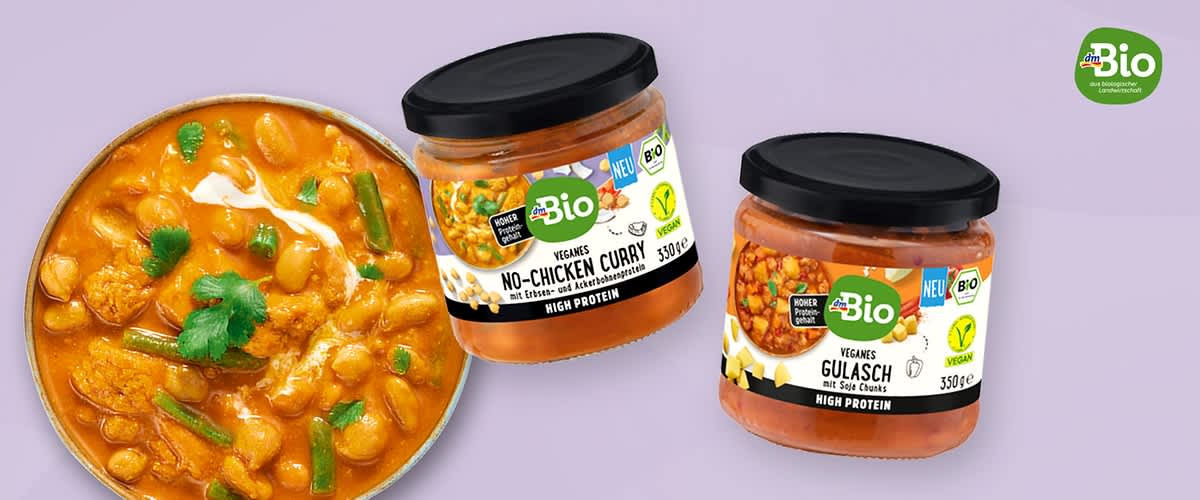 Zwei Glasgläser mit dmBio-Aufklebern, links ein Glas mit der Aufschrift „No‑Chicken Curry“ und rechts ein Glas mit der Aufschrift „Veganes Gulasch“, daneben links eine Schale mit Currysoße und Kichererbsen auf hellem violettem Hintergrund