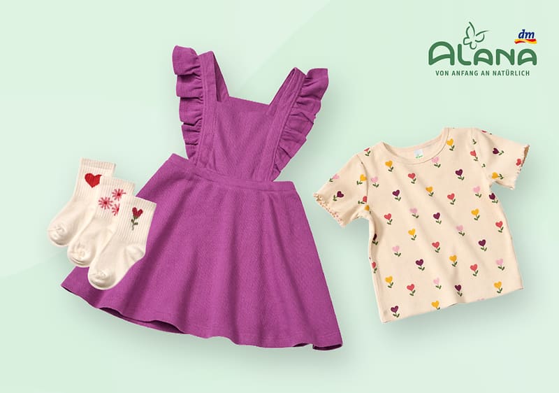Ein lila Kinderkleid mit Rüschenärmeln, daneben ein cremefarbenes T-Shirt mit bunten Blumenmotiven. Links sind drei Paar weiße Socken mit Herz- und Blumenmustern. Oben rechts das Logo von Alana und dm.