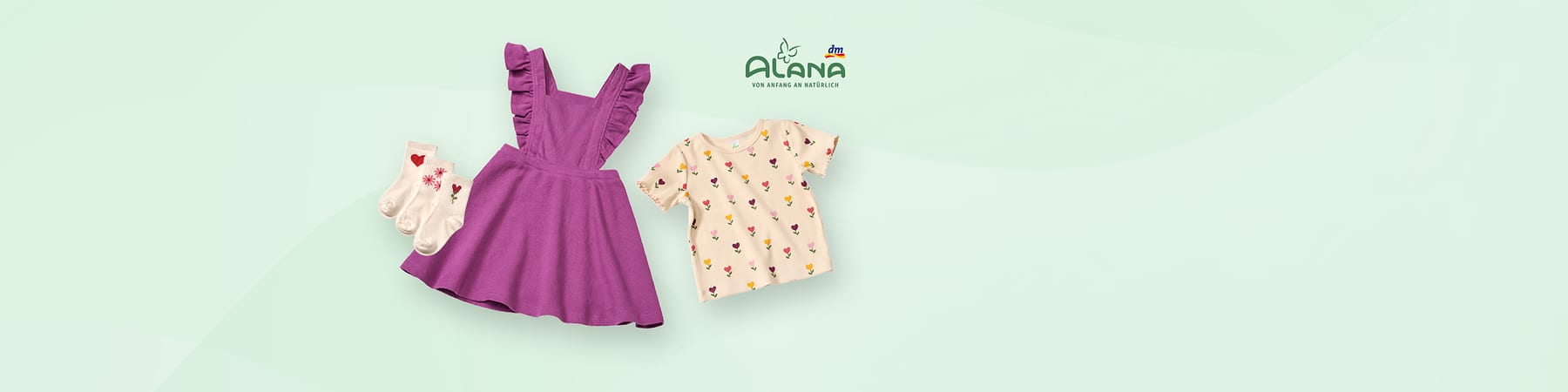 Ein lila Kinderkleid mit Rüschenärmeln, daneben ein cremefarbenes T-Shirt mit bunten Blumenmotiven. Links sind drei Paar weiße Socken mit Herz- und Blumenmustern. Oben rechts das Logo von Alana und dm.