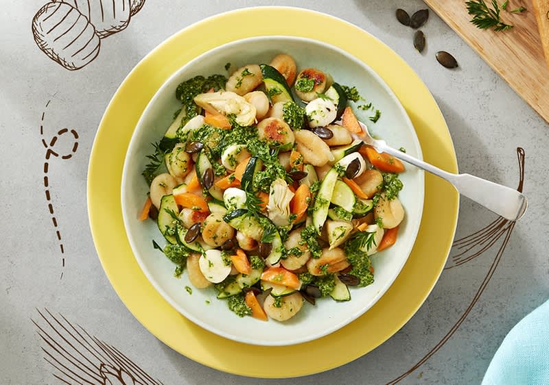 Ein Teller mit einem gemischten Salat aus Gnocchi, Zucchini, Karotten und Kräutern, garniert mit Kürbiskernen, auf einem gelben Unterteller mit einer Gabel rechts daneben