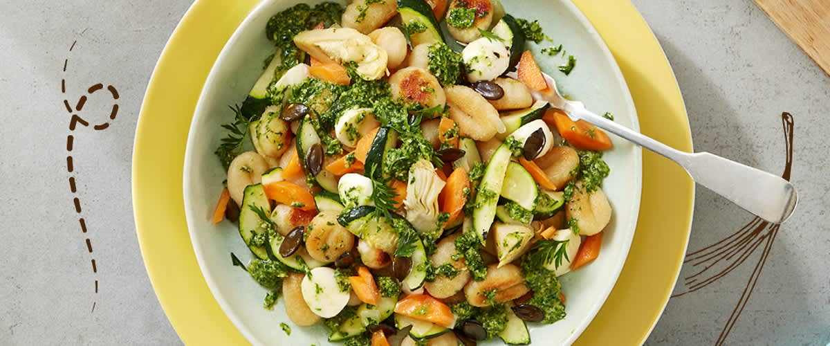 Ein Teller mit einem gemischten Salat aus Gnocchi, Zucchini, Karotten und Kräutern, garniert mit Kürbiskernen, auf einem gelben Unterteller mit einer Gabel rechts daneben