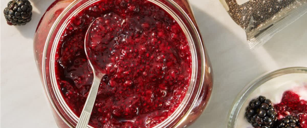 Ein Glas mit roter Beerenmarmelade und einem Löffel darin, daneben ein Glas mit Joghurt und Brombeeren sowie eine Packung Chiasamen