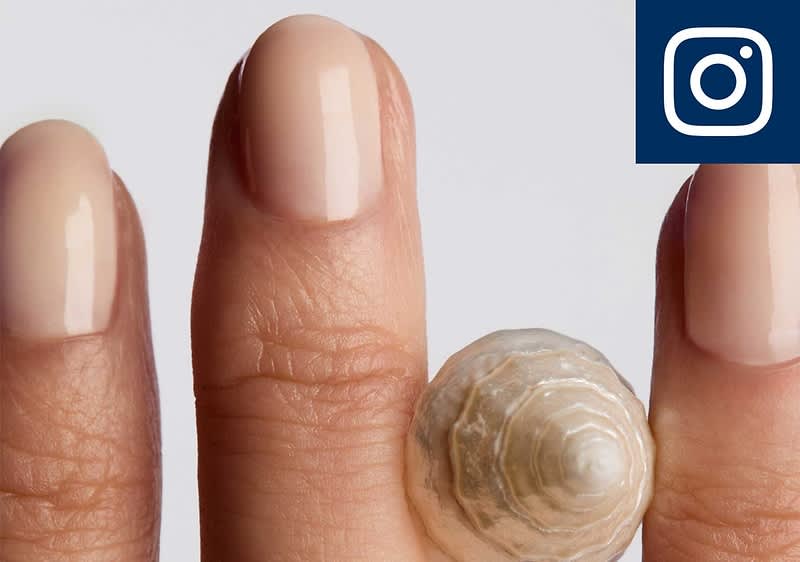 Zwei Finger mit hellrosa lackierten Nägeln, zwischen ihnen auf dem Mittelfinger eine kleine, beige-weiß gestreifte Schneckenhaus-Perle und oben rechts das Instagram-Logo