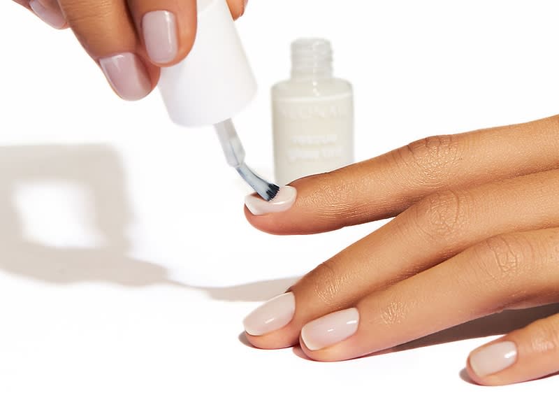 Helle Hände mit kurzen, gepflegten Nägeln, eine Hand trägt hellrosa Nagellack, die andere Hand hält einen weißen Nagellackpinsel und lackiert den Ringfinger, im Hintergrund eine offene weiße Nagellackflasche ohne lesbaren Text
