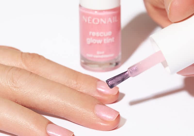 Eine Hand mit rosa lackierten Nägeln wird mit einem rosa Nagellack von NEONAIL „rescue glow tint“ lackiert, der im Hintergrund steht