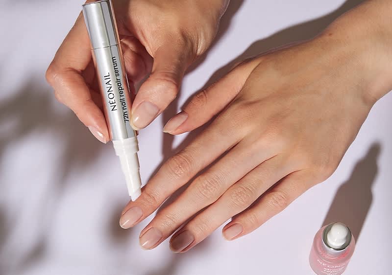 Zwei Hände mit gepflegten, natürlichen Nägeln, die rechte Hand liegt flach auf einer hellen Oberfläche, die linke Hand hält einen silbernen Nagelpflege-Stift mit der Aufschrift „NEONAIL treatment cuticle remover“ und trägt damit Produkt an der Nagelhaut des Mittelfingers auf, neben den Händen eine geöffnete kleine rosa Flasche mit silberner Kappe