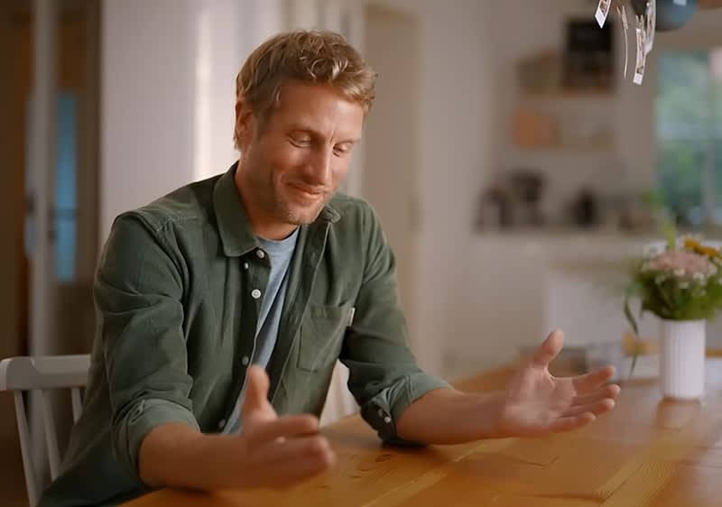 Ein Mann in einem grünen Hemd sitzt an einem Holztisch und gestikuliert mit den Händen. Im Hintergrund sind unscharfe Regale und eine Vase mit Blumen sichtbar.