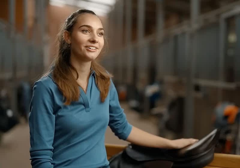 Eine Person in einem blauen Pullover sitzt in einem Stall und hält einen Sattel in der Hand