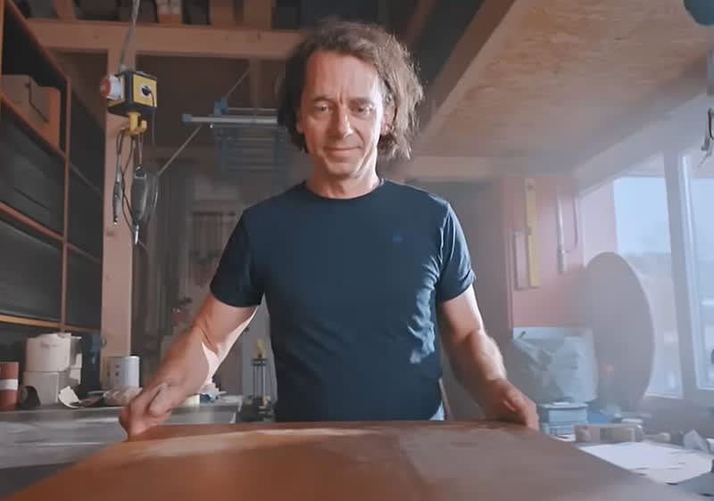 Eine Person in einem blauen T-Shirt steht in einer Werkstatt und hält ein Holzbrett in den Händen. Im Hintergrund sind Regale und Werkzeuge zu sehen.