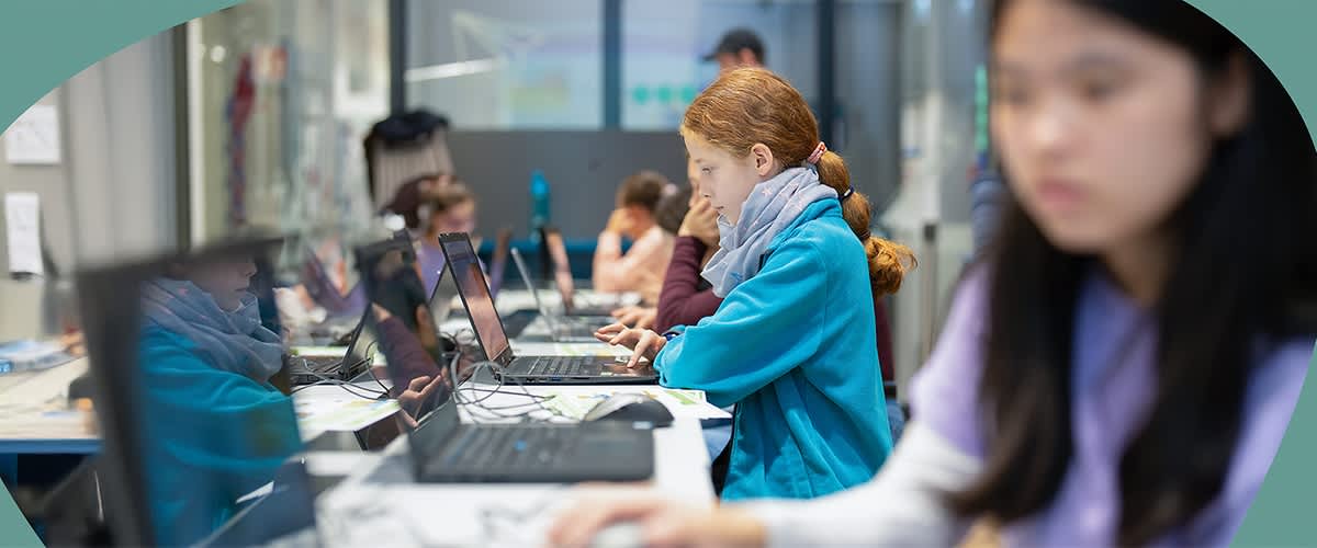 Mehrere Personen arbeiten konzentriert an Laptops in einem modernen Büro. Eine Person im Vordergrund trägt einen blauen Pullover und einen Schal.