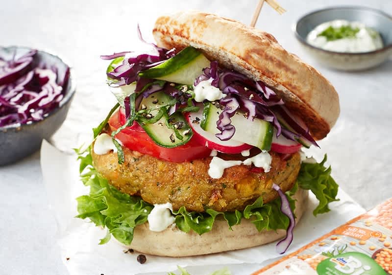 Ein vegetarischer Burger mit einem Linsen-Patty, frischem Salat, Tomatenscheiben, Gurken, Rotkohl und einer weißen Sauce auf einem Brötchen. Im Hintergrund eine Schale mit Rotkohl und ein kleiner Teller mit Sauce