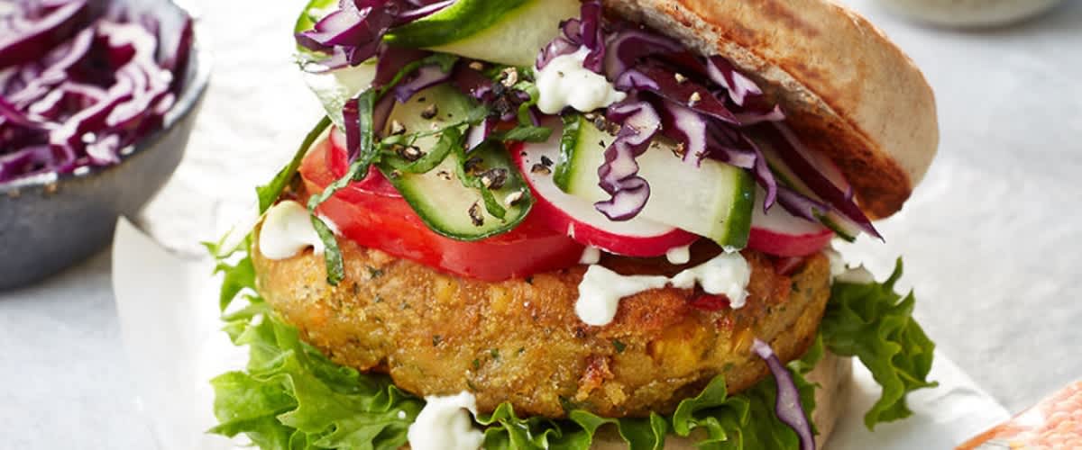 Ein vegetarischer Burger mit einem Linsen-Patty, frischem Salat, Tomatenscheiben, Gurken, Rotkohl und einer weißen Sauce auf einem Brötchen. Im Hintergrund eine Schale mit Rotkohl und ein kleiner Teller mit Sauce
