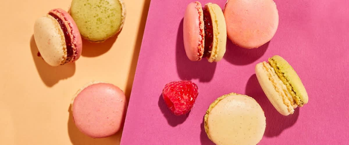 Macarons in verschiedenen Farben liegen auf einem orangefarbenen und einem pinken Hintergrund, begleitet von Himbeeren, einer gezeichneten Mandel und einem gezeichneten Marmeladenglas.