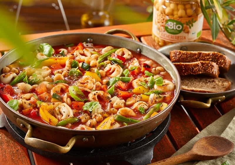 Ein Pfannengericht mit Tortellini, buntem Gemüse und Kichererbsen in Tomatensauce, daneben eine Scheibe Vollkornbrot und ein Glas Bio-Kichererbsen