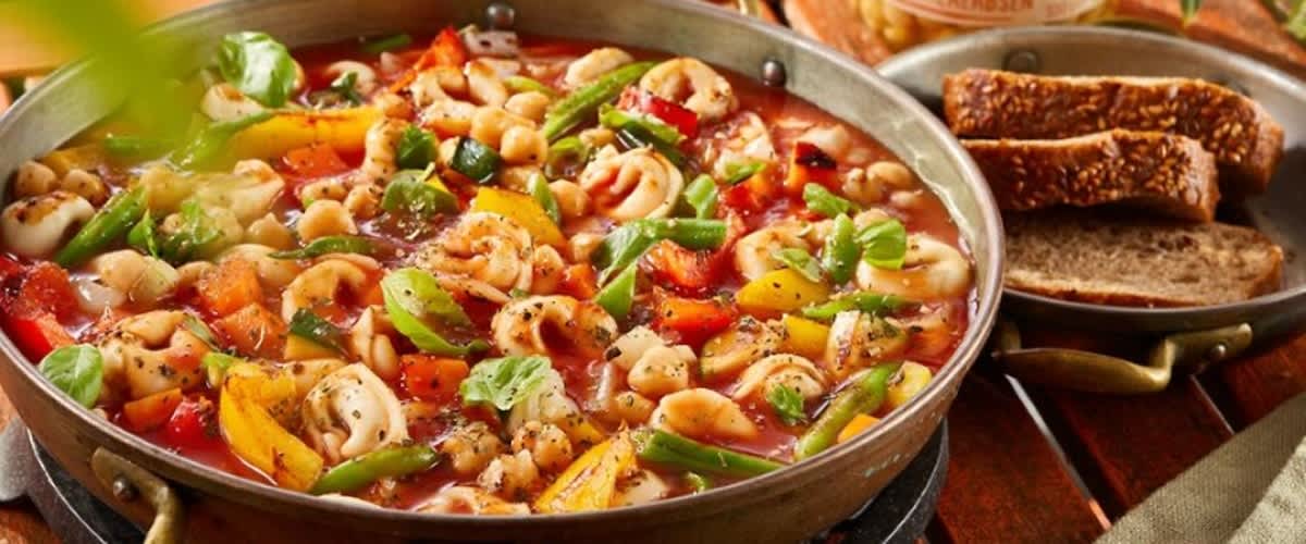 Ein Pfannengericht mit Tortellini, buntem Gemüse und Kichererbsen in Tomatensauce, daneben eine Scheibe Vollkornbrot und ein Glas Bio-Kichererbsen