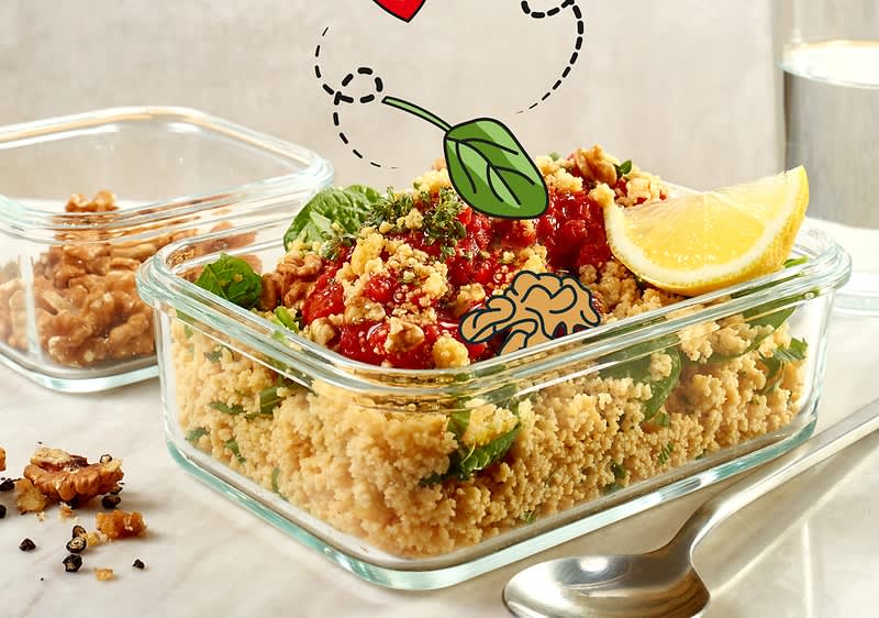 Ein Glasbehälter mit Couscous, garniert mit Tomatensauce, frischen Kräutern und einer Zitronenscheibe. Daneben eine Schale mit Walnüssen und ein Glas Wasser. Illustrationen von einem Herz und einem Basilikumblatt sind über dem Couscous platziert.